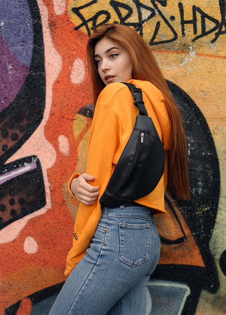 May include: A black fanny pack with a zippered pocket and adjustable strap. The bag is worn across the body. The person is wearing an orange hoodie and blue jeans. The background is a colorful graffiti wall.