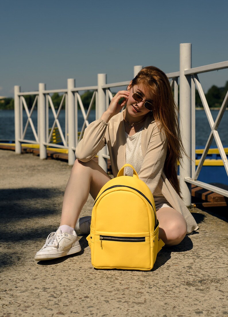 May include: A bright yellow backpack with a black zipper and trim sits on a concrete surface. The backpack is the focal point, with a person and a waterfront background. The bag has a top handle and a front zippered pocket.