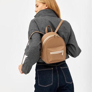 Könnte beinhalten: Ein kleiner, beigefarbener Rucksack mit glatter, matter Oberfläche. Die Tasche verfügt über ein Hauptfach mit Reißverschluss und weißem Rand, eine kleinere Reißverschlusstasche auf der Vorderseite, einen Tragegriff und dünne, verstellbare Schultergurte.