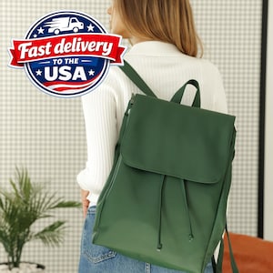May include: A dark green backpack featuring a fold-over flap closure with a drawstring mechanism. The bag has adjustable shoulder straps and a top handle, constructed from a smooth, solid-coloured material with a clean, structured design.