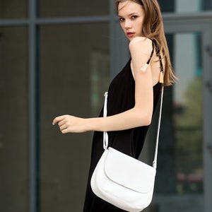 Pu&ograve; includere: Una donna indossa un abito nero e una borsa bianca a tracolla con patta curva. La borsa ha una tracolla lunga e sottile. L'abito ha spalline sottili con nappe decorative. Lo sfondo &egrave; un edificio moderno.