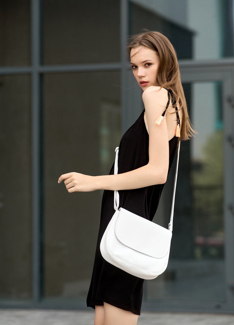 Pu&ograve; includere: Una donna indossa un abito nero e una borsa bianca a tracolla con patta curva. La borsa ha una tracolla lunga e sottile. L'abito ha spalline sottili con nappe decorative. Lo sfondo &egrave; un edificio moderno.