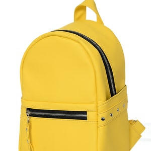 May include: A bright yellow backpack with a rounded top and a small handle. The backpack has a black zipper and a front pocket with a zipper. Silver studs are on the side of the backpack.