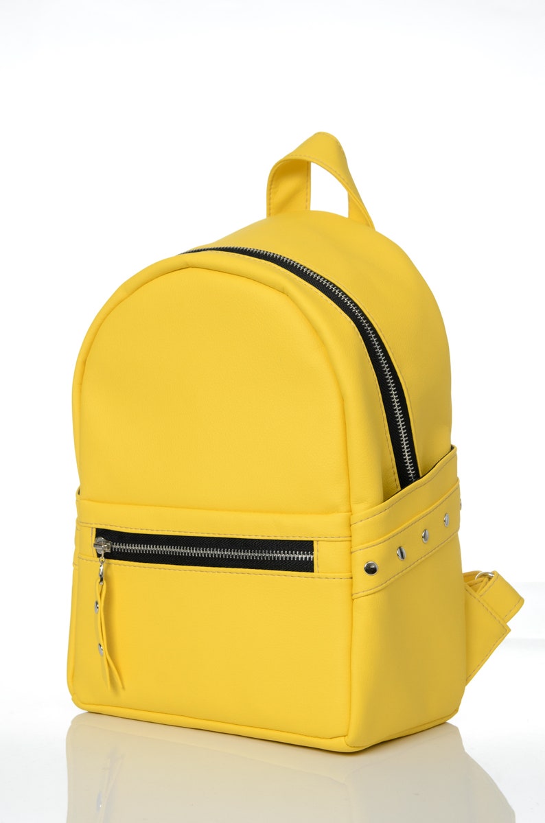 May include: A bright yellow backpack with a rounded top and a small handle. The backpack has a black zipper and a front pocket with a zipper. Silver studs are on the side of the backpack.