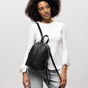 May include: A black backpack featuring a woven leather-like texture, a vertical zippered front pocket, and thin adjustable shoulder straps. The bag has a rounded top silhouette with a small handle at the top and a central zipper closure.