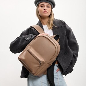 May include: A taupe-colored backpack featuring a smooth exterior, a front zippered pocket with a silver-toned metal zipper, a top handle, and adjustable shoulder straps. The backpack has a rounded top silhouette and a clean, minimalist design.