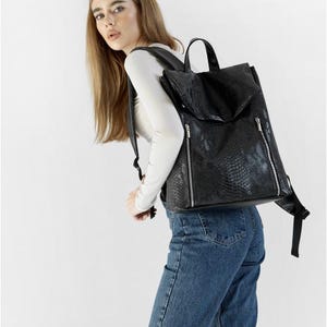 Puede incluir: Mochila negra con un patrón texturizado similar a la piel de serpiente. La mochila tiene un cierre de solapa, dos bolsillos delanteros con cremallera y correas ajustables. La mochila la lleva una persona con una camisa blanca de manga larga y vaqueros azules.