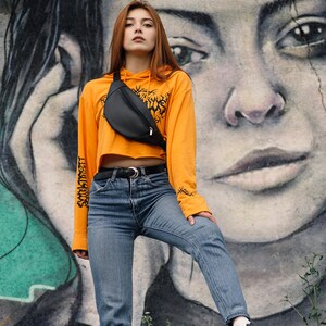 May include: A black fanny pack worn over an orange cropped hoodie and blue jeans. The hoodie has a graphic on the front and text on the sleeves. The background features a large mural of a face.