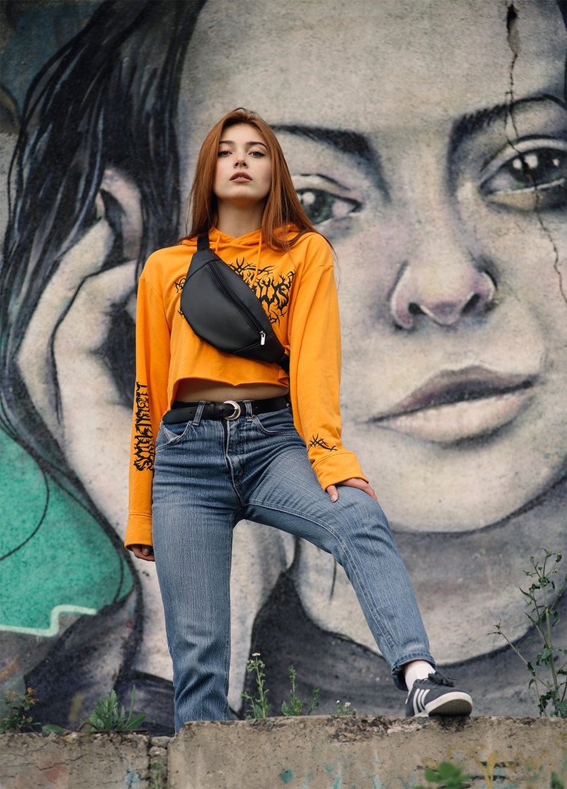 May include: A black fanny pack worn over an orange cropped hoodie and blue jeans. The hoodie has a graphic on the front and text on the sleeves. The background features a large mural of a face.