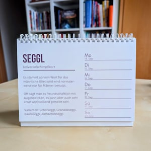 Könnte beinhalten: Ein weißer Schreibtischkalender mit Spiralbindung. Der Kalender zeigt das Wort "SEGGL" in großen lila Buchstaben, darunter deutscher Text. Auf der rechten Seite sind die Daten vom 14. bis 20. September aufgeführt.
