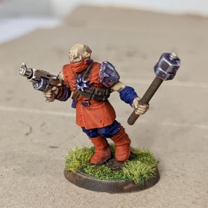 May include: A painted miniature figure of a human-like character wearing a red tunic, blue pants, and a purple and blue shoulder pad. The figure is holding a gun in one hand and a metal club in the other. The figure is standing on a green base.