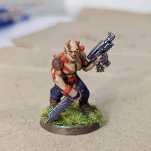 May include: A painted miniature figure of a human-like creature with red and white paint on its torso, blue pants, and a purple gun. The figure is holding a blue sword and standing on a green base.