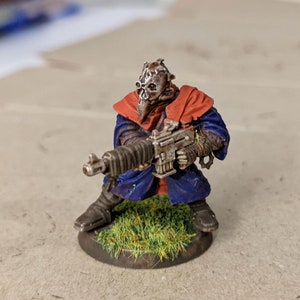May include: A miniature figure of a person wearing a blue and red robe, holding a gun. The figure is standing on a base covered in green grass.
