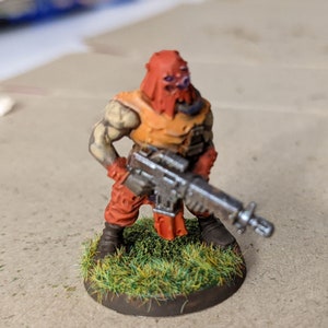 May include: A miniature figure of a soldier wearing a red and orange hooded outfit, holding a silver rifle. The figure is standing on a green grass base.