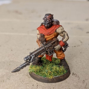 May include: A painted miniature figure of a sci-fi soldier wearing a red and orange jumpsuit and a helmet with a gas mask. The figure is holding a black and silver gun and is standing on a green base.