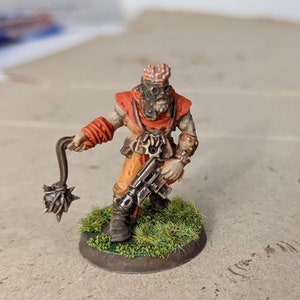 May include: A painted miniature figure of a human-like character with a spiked club, wearing a red and orange outfit, holding a gun and standing on a green base.