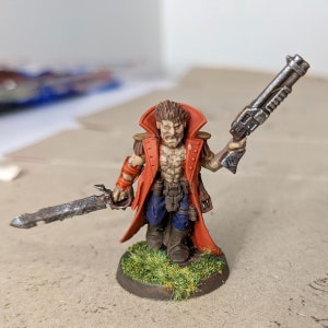 May include: A red-coated miniature figure with a sword and a gun, standing on a green base. The figure has a detailed face and a scarred chest.
