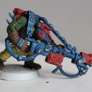 Könnte beinhalten: Eine grün und gelb bemalte Miniaturfigur eines Orks, der eine blaue und rote Waffe mit goldenen Akzenten hält. Die Figur steht auf einem grauen Sockel.