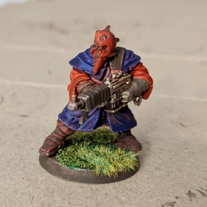 May include: A red and blue painted miniature figure of a person wearing a mask and holding a gun. The figure is standing on a green base.