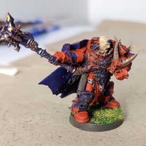 May include: A miniature figurine of a warrior in red and blue armor, holding a gold and purple weapon. The figure has a skull-like head and is standing on a green, grassy base. The figure is a fantasy character.