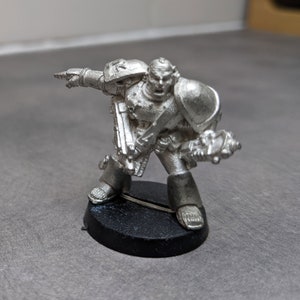 Peut inclure: Une figurine miniature en métal argenté représentant un soldat de l'espace marine portant une armure et tenant une arme. La figurine est posée sur une base noire.