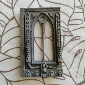 Peut inclure: Un cadre de fenêtre en plastique gris avec un design en arc gothique. Le cadre est décoré de crânes et de chaînes. La fenêtre a un motif de grille.