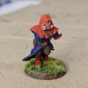 May include: A painted miniature figure of a person wearing a red hooded robe and a gas mask. The figure is standing on a green grassy base.
