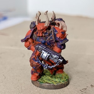 May include: A red and blue painted miniature figure of a space marine with a horned helmet and a chain-linked weapon. The figure is standing on a green base.