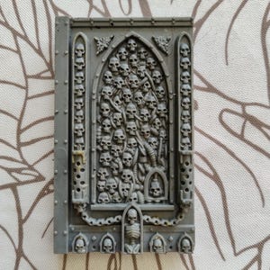 Peut inclure: Un modèle en plastique gris d'une arche gothique avec un motif de crâne. L'arche est remplie de crânes et d'os, et il y a des chaînes et d'autres détails.