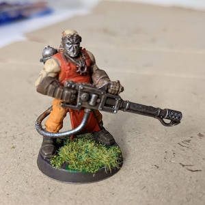 May include: A painted miniature figure of a sci-fi soldier holding a large gun. The figure is wearing a red and orange jumpsuit and a metal mask. The figure is standing on a green base.