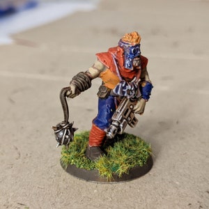 May include: A miniature figure of a warrior with a spiked chain weapon and a gun. The figure is wearing a red, orange, and blue outfit and a mask. The figure is standing on a green base.