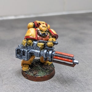 May include: A yellow and red painted miniature space marine figure holding a silver and red gun. The figure is standing on a brown base with green and brown painted details.