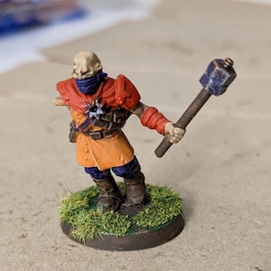 May include: A miniature figure of a person wearing a red and orange outfit with a blue scarf and holding a blue and brown weapon. The figure is standing on a green base.