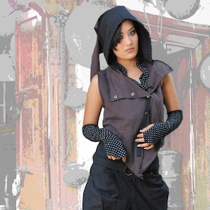 Könnte beinhalten: Eine Frau trägt eine braune Kapuzenweste mit schwarzen und weißen Polka-Dot-Ärmeln, schwarze Hosen und braune Stiefel. Sie steht vor einer Wand mit einer Holztür und verschiedenen Metallgegenständen, die von der Decke hängen.