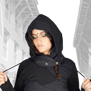 Könnte beinhalten: Ein schwarzer Kapuzenpullover mit einem geschichteten Design und einem schwarzen Gürtel mit goldenen Akzenten. Der Pullover hat lange Ärmel und einen hohen Kragen. Das Model trägt den Pullover in einer Straßenumgebung.