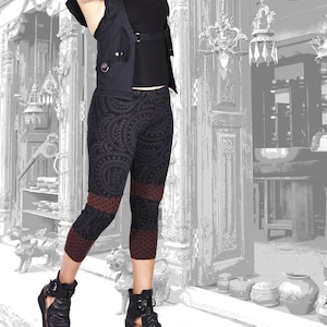 May include: A woman wearing a black hooded vest with a zipper and pockets over a black tank top and black and brown patterned leggings. She is standing in front of a doorway with ornate details and hanging light fixtures.