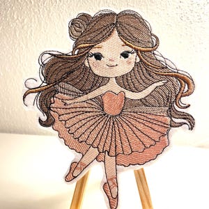 Könnte beinhalten: Gestickter Aufnäher einer Ballerina mit braunen Haaren, die ein rosafarbenes Tutu und rosafarbene Ballettschuhe trägt. Der Aufnäher befindet sich auf einem kleinen Holzstaffelei.