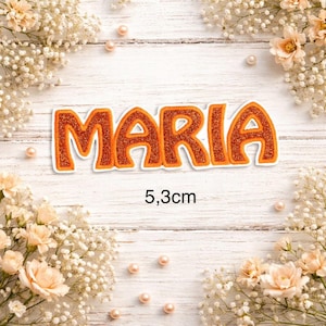 Könnte beinhalten: Ein glitzernder, orangefarbener Aufbügler mit dem Namen MARIA in einer fetten, abgerundeten Schrift mit weißem Rand. Der Text ist über der Maßangabe 5,3 cm zentriert, die in Schwarz auf einem weißen Holzmaserungshintergrund mit Blumen abgebildet ist.