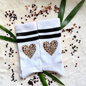 Könnte beinhalten: Ein Paar weiße Socken mit zwei horizontalen schwarzen Streifen am oberen Rand und einem herzförmigen Leopardenmuster in der Mitte jeder Socke. Die Socken sind gerippt und aus weißem Stoff gefertigt, was einen deutlichen Kontrast erzeugt.