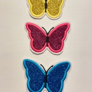Könnte beinhalten: Drei glitzernde Schmetterlings-Aufnäher zum Aufbügeln in Gelb, Pink und Blau.