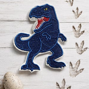 Könnte beinhalten: Ein dunkelblauer, glitzernder T-Rex-Dinosaurier-Aufnäher mit weißem Rand, rotem Maul, spitzen weißen Zähnen und einem gelben Auge. Der Aufnäher wird zusammen mit einem gesprenkelten Ei und mehreren Dinosaurier-Fußabdrücken dargestellt.