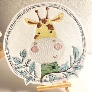 Könnte beinhalten: Gestickte Filzapplikation einer Giraffe mit einem grünen Shirt und einem Herz darauf. Die Giraffe ist von blauen und grünen Blättern in einem runden Rahmen umgeben.