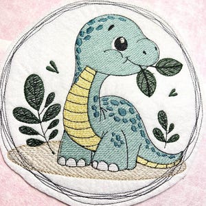Könnte beinhalten: Stickerei mit einem niedlichen grünen Dinosaurier mit gelben Streifen, der ein Blatt frisst. Der Dinosaurier ist von einem Kreis aus Blättern und Herzen umgeben.