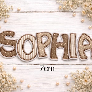 Könnte beinhalten: Der Name SOPHIA, geschrieben in einzelnen Stoffbuchstaben. Die Buchstaben wechseln zwischen einem braunen Stoff mit weißen Punkten und einem beige-weiß gestreiften Stoff. Jeder Buchstabe hat einen weißen Rand und ist 7 cm hoch.