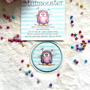 Könnte beinhalten: Eine quadratische Karte und ein runder Aufnäher mit einer Illustration eines rosa-violetten Monsters auf hellblau gestreiftem Grund. Die Karte enthält den Text Mutmonster und ein deutsches Gedicht. Kleine bunte Metallglöckchen liegen verstreut.