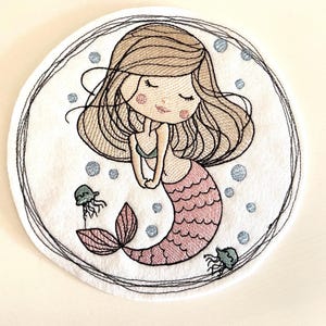 Könnte beinhalten: Bestickter weißer Untersetzer mit einer Cartoon-Meerjungfrau mit braunem Haar, einem rosa Schwanz und einem blauen Bikini-Oberteil. Das Design umfasst zwei Quallen und blaue Blasen, die alle von einem schwarzen, genähten Rand umgeben sind. Ein skurriles, maritimes Thema.