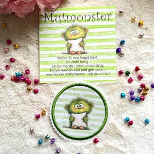 Könnte beinhalten: Eine quadratische Karte mit grün-weißen Querstreifen, einer grünen Monsterillustration und dem Text Mutmonster, dazu ein passender runder, bestickter Aufnäher mit gezacktem Rand und grünem Saum.