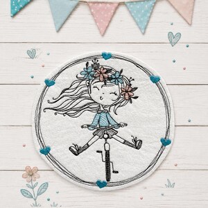 Könnte beinhalten: Ein runder, weißer Stoffaufnäher mit einer gestickten Illustration einer Person mit langem Haar und Blumenkranz auf einem Fahrrad. Das Motiv ist von einem dünnen, schwarzen Kreis umgeben, der mit kleinen blauen Stickherzen verziert ist.