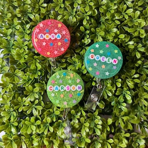 May include: Three glittery badge reels with star accents. One is red with the name "ANNA", another is green with "QUEEN", and the last is green with "CARSON". Each reel has a clear plastic clip and a retractable cord.