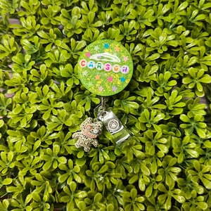 May include: A green glitter badge reel with the name "CARSON" in colourful letters. The reel has a small charm of a leopard-print creature. The background is a bed of green leaves.